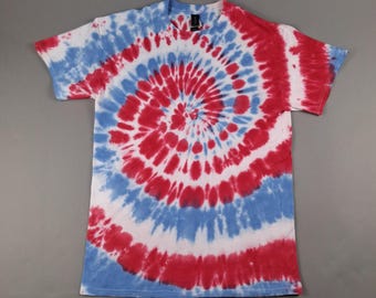 May include: A short-sleeved t-shirt with a red, white, and blue spiral tie-dye pattern. The design radiates from the center of the shirt, creating a circular effect. The shirt is made of a soft material and has a crew neck.