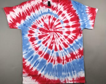 May include: A red, white, and blue tie-dye t-shirt. The design features a spiral pattern with red and white radiating from the center, surrounded by a blue band. The shirt has short sleeves and a crew neck.