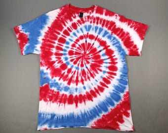 May include: A red, white, and blue tie-dye t-shirt. The spiral design features concentric circles of red, white, and blue. The shirt has a crew neck and short sleeves. The shirt is made of a soft material.