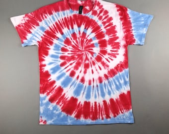 May include: A short-sleeved t-shirt with a spiral tie-dye pattern in red, white, and blue. The design radiates from the center, creating a bold and colorful effect. The shirt is made of a soft material and is suitable for casual wear.