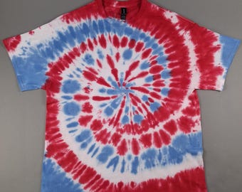 May include: A short-sleeved t-shirt with a spiral tie-dye pattern in red, white, and blue. The design radiates from the center, with alternating bands of color. The shirt is laid flat on a gray surface.