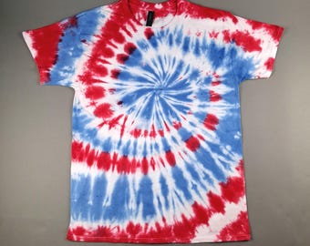 Könnte beinhalten: Ein rot, weiß und blaues Batik-T-Shirt. Das Spiraldesign weist konzentrische Kreise in Rot, Weiß und Blau auf. Das Shirt hat kurze Ärmel und einen Rundhalsausschnitt. Das Shirt ist aus einem weichen Material gefertigt.