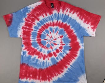 May include: A short-sleeved t-shirt with a spiral tie-dye pattern in red, white, and blue. The design radiates from the center of the shirt, creating a circular pattern. The shirt is laid flat on a gray surface.