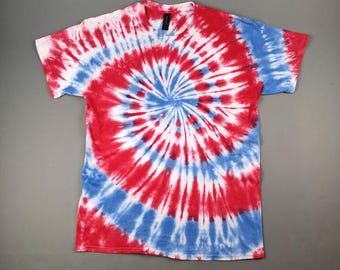 May include: A red, white, and blue tie-dye t-shirt. The spiral design features a central blue circle, surrounded by red and white radiating stripes. The shirt has a V-neck and short sleeves.