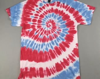 May include: A short-sleeved t-shirt with a spiral tie-dye pattern in red, white, and blue. The design radiates from the center of the shirt, creating a circular effect. The shirt is laid flat on a gray surface.