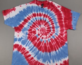 May include: A short-sleeved t-shirt with a spiral tie-dye pattern in red, white, and blue. The design radiates from the center of the shirt, creating a circular pattern. The shirt is laid flat on a gray surface.