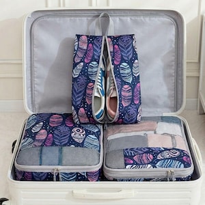 Op de afbeelding: Een witte koffer is open en toont reisorganizers. De set bevat een schoenentas met sneakers en inpakblokjes. Het verenpatroon heeft marineblauwe, paarse, roze en turquoise accenten.