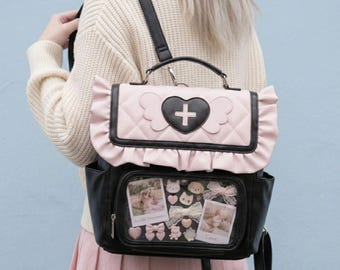 以下が含まれることがあります： 黒とライトピンクのバックパック。キルティング加工されたフラップには、白い十字架が入った黒いハートと翼のエンブレムが付いています。バッグの前面には透明なディスプレイウィンドウがあり、小さな装飾ピン、リボン、2枚の長方形の写真が入っています。
