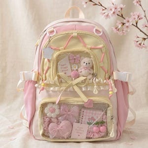 Puede incluir: Una mochila de color rosa pastel y crema con dos bolsillos frontales transparentes llenos de artículos decorativos, incluyendo un pequeño oso de peluche, un lazo de cuadros rosa, un cuaderno con ilustraciones de tulipanes y tarjetas que dicen Good Things Ahead y Enjoy the little things.