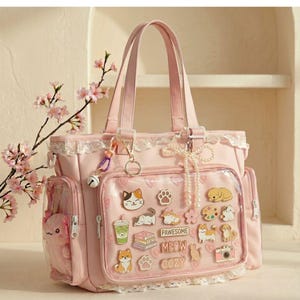 May include: A pale pink tote bag featuring a clear front pocket decorated with cat and dog-themed pins. The bag has lace trim, a pearl charm, and a small bell. A plush toy is visible in a side pocket. The bag has two handles.