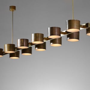 Könnte beinhalten: Eine moderne, messingfarbene Hängeleuchte mit zylindrischen Schirmen. Die Leuchte hat ein lineares Design mit mehreren Lichtern, die an einer zentralen Stange aufgehängt sind. Die Schirme sind gleichmäßig verteilt und strahlen ein warmes Licht aus.