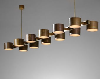May include: A modern, brass-toned chandelier with cylindrical shades. The fixture has a linear design with multiple lights, suspended from a central rod. The shades are evenly spaced and emit a warm glow.