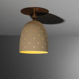 Könnte beinhalten: Eine beige, glockenförmige Deckenleuchte mit kleinen, gleichmäßig verteilten Löchern. Die Leuchte hat eine bronzefarbene Oberseite und einen Stiel. Das Licht wird von innen beleuchtet und wirft einen warmen Schein.