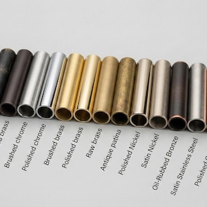 May include: A collection of fifteen cylindrical metal samples arranged in a row, each featuring a distinct finish labeled as matte black, antique black, blackened brass, brushed chrome, polished chrome, brushed brass, polished brass, raw brass, antique patina, polished nickel, satin nickel, oil-rubbed bronze, satin stainless steel, polished copper, and aged verdigris.