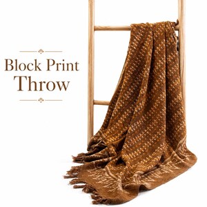 May include: A brown throw blanket with a repeating block print pattern of small geometric shapes and a fringed edge, draped over a light-colored wooden ladder. The text Block Print Throw is printed in brown on the left side of the image.