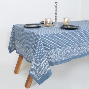 May include: A blue tablecloth with a white leaf pattern and a decorative border. The tablecloth is set on a wooden table with black plates, gold-rimmed glasses, and a silver candlestick holder. The table is set for a meal.