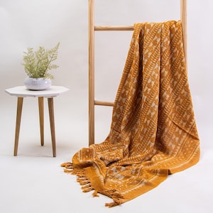 Op de afbeelding: Een mosterdgele plaid met een wit varenachtig botanisch patroon en franjes aan de randen, gedrapeerd over een lichthouten ladder. Een kleine witte pot met een varen staat op een wit bijzettafeltje met drie poten naast de ladder.