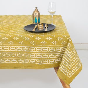 May include: A mustard yellow tablecloth with a white geometric pattern. A black plate with gold cutlery, a gold napkin ring, and a decorative lantern are on the table. A wine glass is also present.