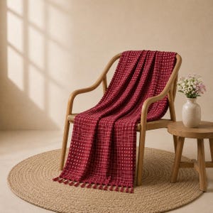 Puede incluir: Una manta de color rojo intenso con un patrón de cuadrícula blanca y flecos, colocada sobre una silla de madera clara. La manta presenta un tejido texturizado con pequeños cuadrados blancos y finas líneas que se cruzan en la tela.