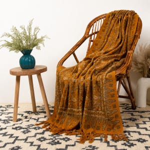 May include: A mustard yellow throw blanket with a dark brown intricate botanical and geometric pattern, featuring a fringed edge, draped over a brown rattan chair. The blanket displays a repeating design of leaves and small square motifs.