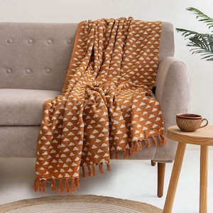 May include: A rectangular throw blanket in a deep burnt orange color featuring a repeating pattern of small white triangles. The blanket has a textured fabric finish and is finished with short, matching orange fringe along the bottom edge.