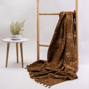 May include: A brown throw blanket featuring a repeating floral pattern and fringe trim along the edges. The fabric displays a textured weave with lighter brown and white floral motifs throughout the design, draped over a wooden ladder frame.