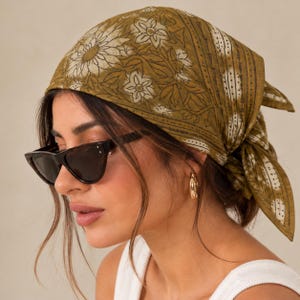 Puede incluir: Un pañuelo cuadrado de color verde oliva con un estampado floral blanco y un borde con líneas punteadas. La tela está anudada en la parte posterior de la cabeza. El accesorio se combina con gafas de sol negras de ojo de gato y un pendiente de aro dorado.