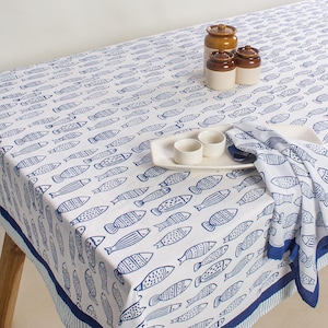 Op de afbeelding: Wit tafelkleed met een blauw vispatroon. Het tafelkleed ligt op een houten tafel en heeft een blauwe rand. Een bijpassende servet en een serveerschaal met twee kleine witte kommen staan ook op de tafel.
