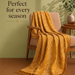 Pode incluir: Uma manta amarelo-mostarda com um padrão branco de ziguezague e pontos, com bordas de franjas. A manta está sobre uma cadeira de madeira. O texto "Perfect for every season" com um pequeno ícone de coração é visível na parede acima da cadeira.