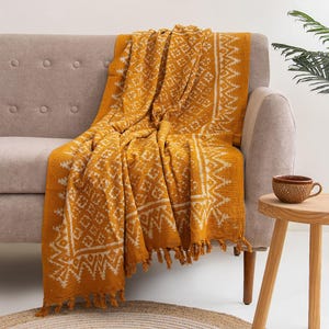 Puede incluir: Una manta de color amarillo mostaza con un patrón geométrico blanco que presenta un diseño de borde repetitivo de diamantes y zigzags. El tejido rectangular tiene una textura marcada y está rematado con flecos cortos y anudados.