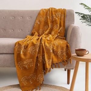 May include: A mustard yellow throw blanket featuring a repeating white tree pattern and fringed edges. The fabric has a textured, woven appearance, and the blanket is draped over a neutral-colored sofa, showcasing its length and detailed design.
