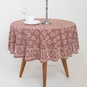 May include: A round wooden table with three tapered legs covered by a dusty rose tablecloth featuring a white floral pattern and a decorative border. On the table sits a stack of white plates, a clear glass wine goblet, and a tall metal candlestick.
