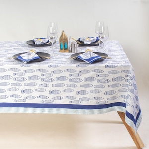 May include: A white tablecloth with a blue fish pattern, set for four. The table is set with black plates, blue napkins, and gold-colored cutlery. A gold lantern and wine glasses complete the nautical theme.