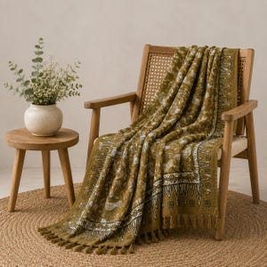 May include: An olive green throw blanket with a white intricate botanical and bird pattern, featuring a decorative border and fringe edges, draped over a wooden chair with a woven cane backrest and a small wooden side table with a vase.