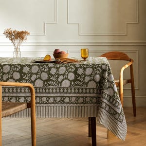 May include: A rectangular tablecloth featuring an olive green base with a white floral pattern, including a detailed border design. The fabric drapes over a table, displaying a repeating botanical motif with stylized flowers and leaves.