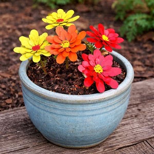 May include: A blue ceramic flower pot filled with soil, showcasing colorful metal flowers. The flowers are yellow, orange, and red with yellow centers. The pot sits on a wooden surface, creating a vibrant garden scene.