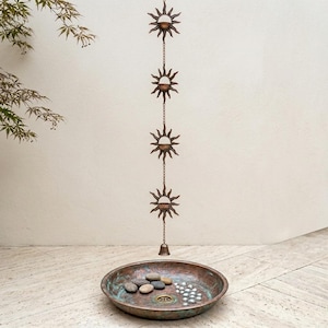 May include: A bronze sun-shaped rain chain with a bell at the bottom, hanging above a copper-colored basin filled with stones and decorative elements. The chain features four sun designs connected by chains, creating a decorative outdoor element.