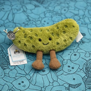 May include: A plush green pickle-shaped toy with a smiling face, small brown legs, and a metal keyring. The toy has a textured surface with darker green spots. A white tag with text is attached to the toy.