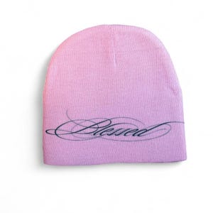 Puede incluir: Un gorro rosa claro con la palabra "Blessed" en escritura cursiva en la parte delantera. El gorro es de color liso y parece estar hecho de un material de punto suave. El texto es negro.
