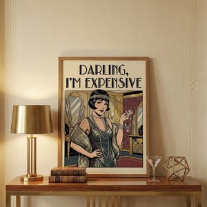 May include: A framed art print featuring a vintage illustration of a woman in a flapper dress holding jewelry. The print has the text "DARLING, I'M EXPENSIVE" in bold black letters. The artwork is displayed on a wooden table with a gold lamp and decorative items.