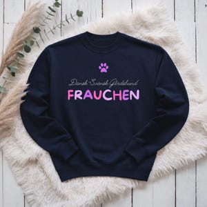 Könnte beinhalten: Marineblaues Sweatshirt mit dem Text "Dansk Svensk Gårdshund FRAUCHEN" in Rosa und Lila. Darüber befindet sich ein lila Pfotenabdruck. Das Sweatshirt liegt auf einer weißen, flauschigen Oberfläche.