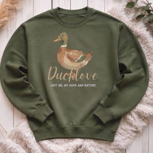 Könnte beinhalten: Olivgrünes Sweatshirt mit einer Entenillustration und dem Text "Ducklove". Darunter steht der Satz "JUST ME, MY DUCK AND NATURE". Das Sweatshirt besteht aus weichem Material und ist für den Freizeitgebrauch geeignet.