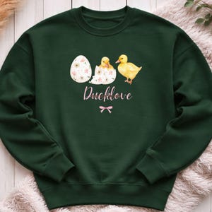 Könnte beinhalten: Ein dunkelgrünes Sweatshirt mit einem Aquarelldesign eines Eis, eines schlüpfenden Entchens und eines gelben Entchens. Der Text "Ducklove" ist in rosa Schrift geschrieben, mit einer rosa Schleife darunter. Das Sweatshirt ist aus weichem Material.