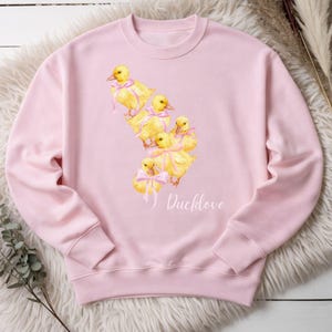 Könnte beinhalten: Hellrosa Sweatshirt mit einem Aquarelldesign von sechs gelben Entenküken mit rosa Schleifen. Das Wort "Ducklove" ist in einer Schreibschrift darunter geschrieben. Das Sweatshirt ist aus einem weichen Material.