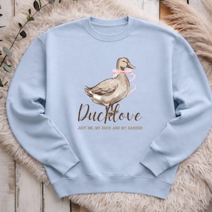 Könnte beinhalten: Hellblaues Sweatshirt mit einer braunen Ente mit rosa Schleife und dem Schriftzug "Ducklove" in brauner Schrift. Darunter steht "Just me, my duck and my garden". Ein lässiger Kleidungsstil.