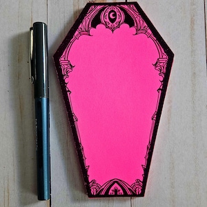 May include: A bright pink coffin-shaped notepad with a black decorative border and a crescent moon design. A dark blue pen is positioned next to the notepad. The notepad is on a light wood surface.