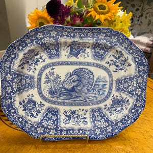 May include: A rectangular ceramic serving platter featuring a blue and white transferware pattern. The center depicts a turkey in a landscape, surrounded by a border of floral motifs, baskets, and star-shaped flowers on a scalloped edge.