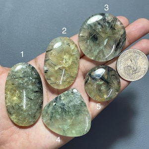 May include: Five polished rutilated quartz cabochons with black needle-like inclusions, labelled 1 through 5, displayed alongside a United States one dollar coin for scale. The stones are translucent with yellow and green hues and smooth surfaces.