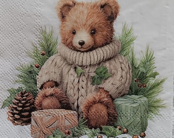 May include: A decorative paper napkin featuring a brown teddy bear wearing a beige sweater and surrounded by pine branches, holly, and wrapped gifts. A pine cone and berries are also present. The napkin has a textured background.