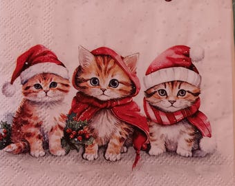 May include: A paper napkin featuring three orange tabby kittens. The left kitten wears a Santa hat, the center kitten wears a red hooded cape, and the right kitten wears a Santa hat and red scarf. A string of colorful Christmas lights borders the top.